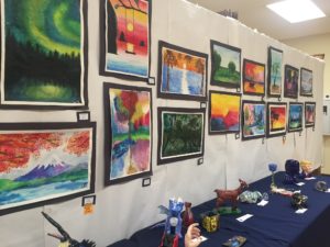 A display at Summit Artspace on Tusc in Barberton of student vibrant watercolor paintings in a display. Below are student ceramic pieces from a cobalt dragon, a lovely fawn to a goblet and other animal figures.