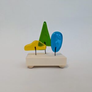 Three abstract glass shapes meant to look like trees in the color yellow, green, and blue, fastened to a light tan wooden block.