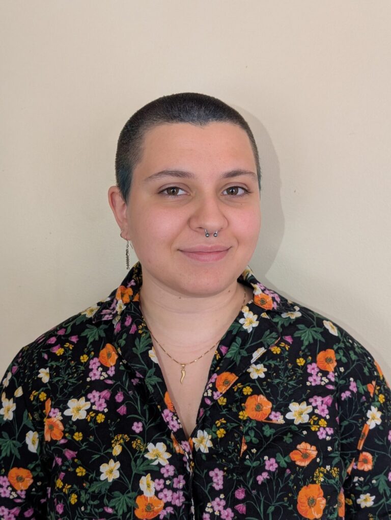 A person with light skin and short buzzed-cut hair wearing a floral printed shirt looks at the camera.