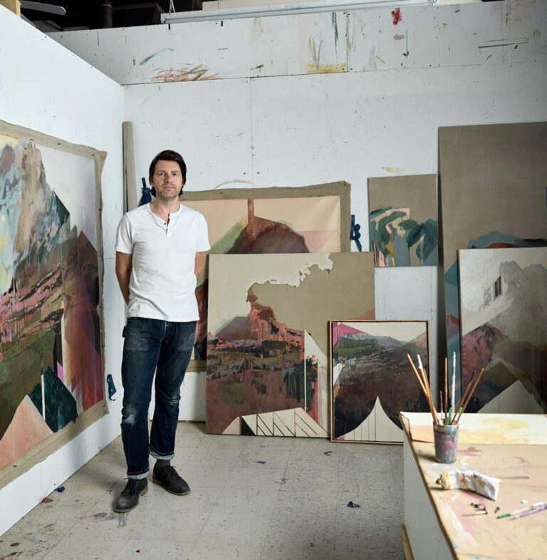 A man with light skin and medium length dark brown hair wearing a white t-shirt and blue jeans stands in front of several abstract paintings.