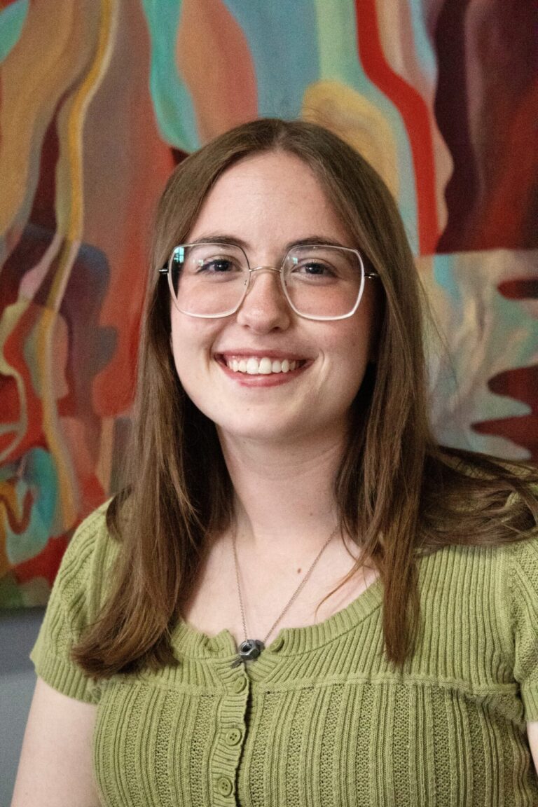 A woman with light skin and long red-brown hair wearing glasses and a green short sleeved t-shirt stands in front of an abstract painting and smiles.