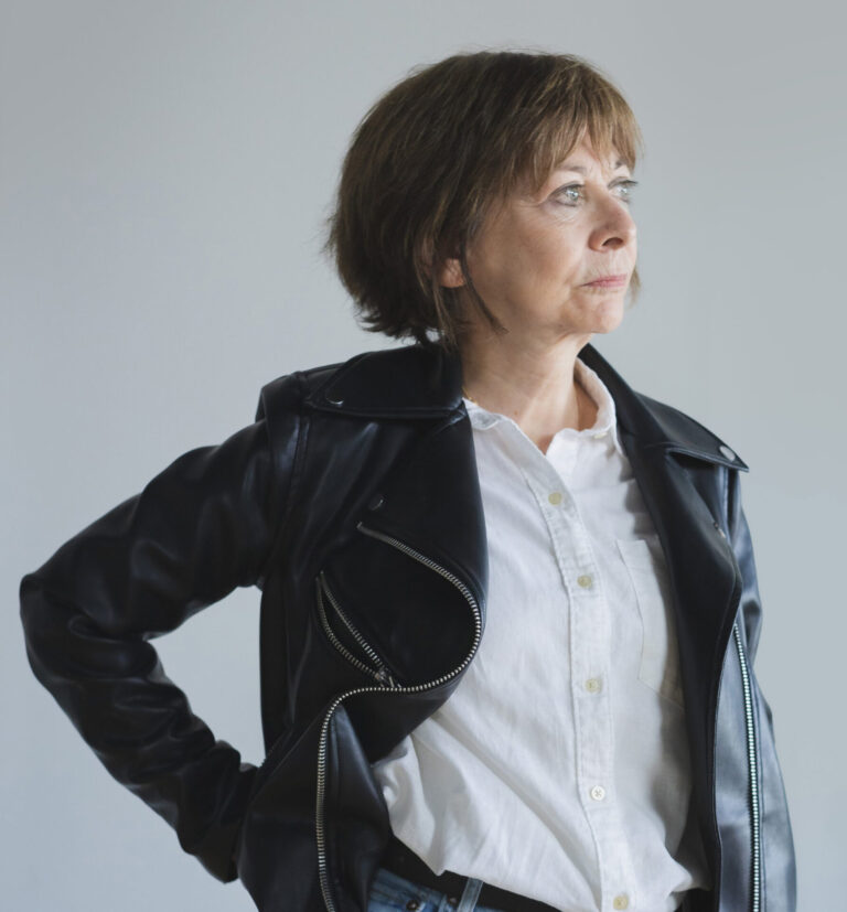 A woman with light skin and a short brown bob haircut, wearing a white shirt and leather jacket looks off to the right side of the camera with her hand on her hip.