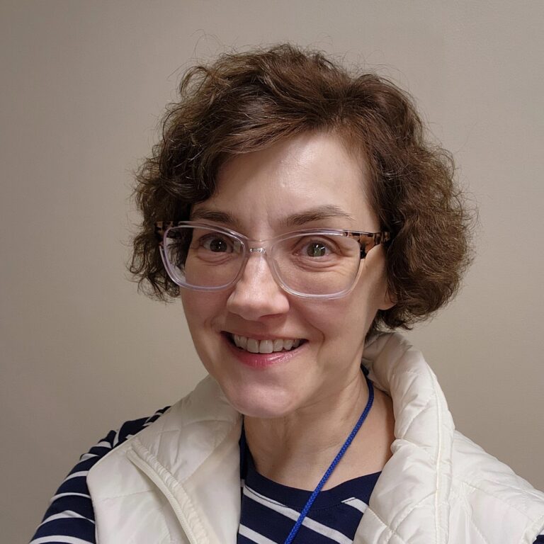 A woman with light skin and a short brown bob haircut parted to one side wearing glasses, a white vest, and a striped shirt underneath smiles at the camera.