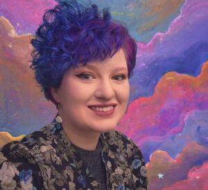 A Person with short blueish purple hair standing and smiling in front of a colorful abstract painting.