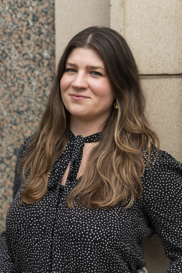 Person with long brown hair with highlights wearing a black patterned blouse smiles at the camera.