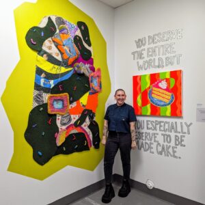 Artist standing and smiling in front of colorful mixed-media wall art and a bright painting of cake with the text 'You deserve the entire world, but you especially deserve to be made cake' on the gallery wall.