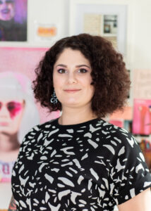 Person with curly dark hair wearing a black and white patterned shirt, standing indoors with colorful artwork blurred in the background.