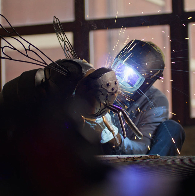 Person welding a creature with wings wearing a welding mask in a dark room.