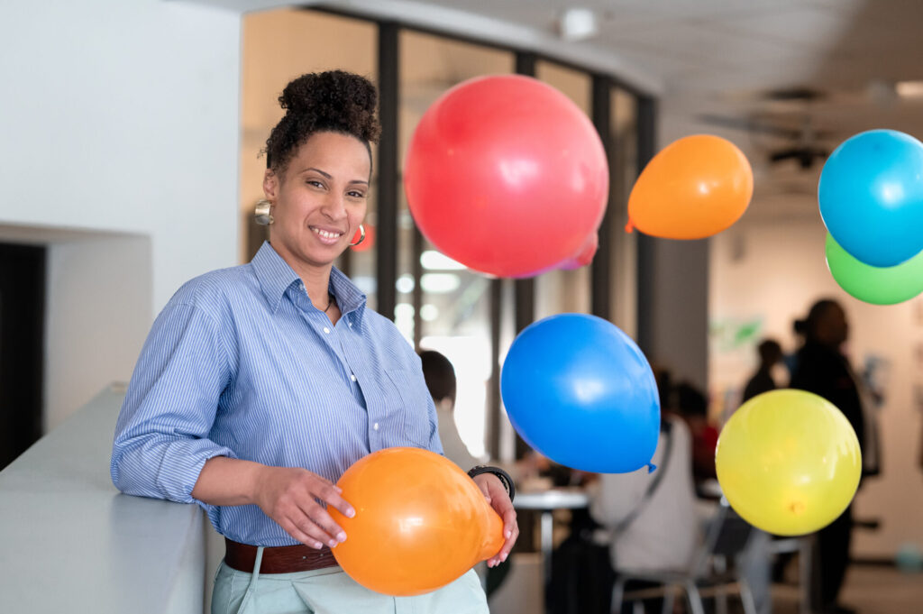 A person stands in a room with multicolored balloons wearing a blue button up shirt and smiles at the camera