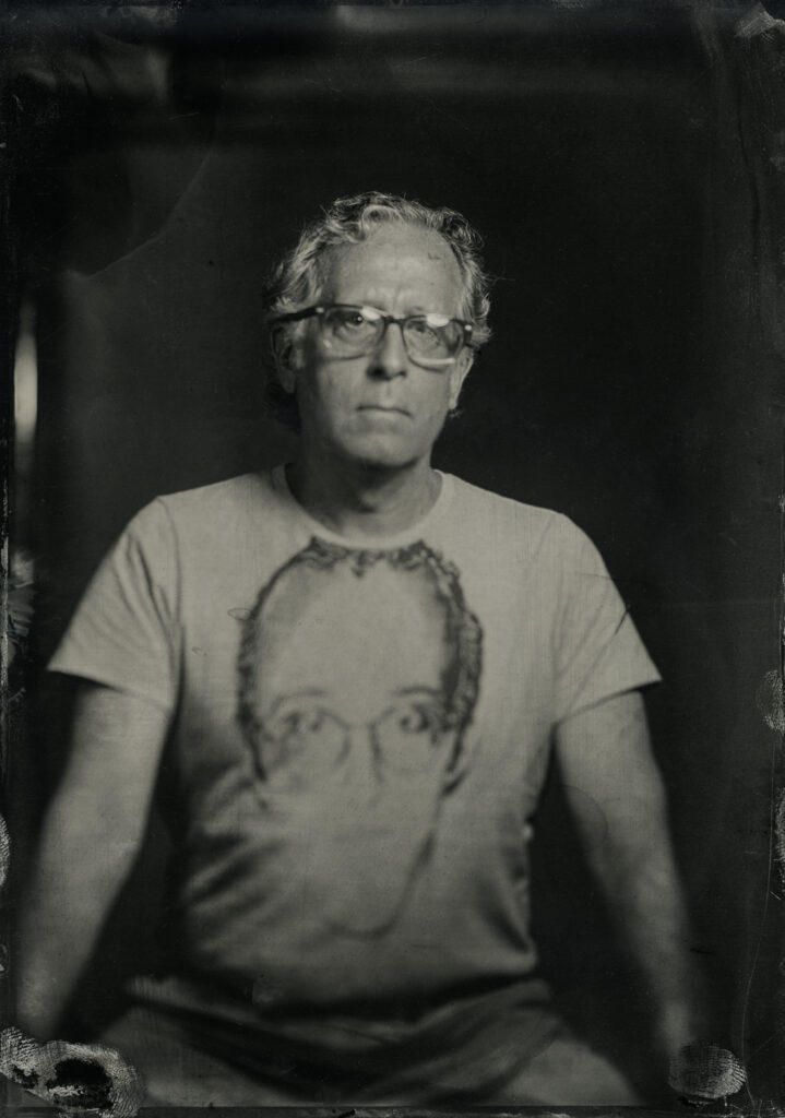 Black and white Tintype of a person wearing a shirt with keith haring on it. The person wearing the shirt is wearing glasses and looks at the camera.