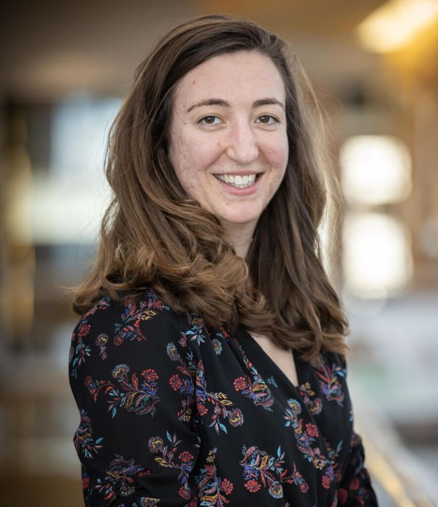 A person with long brown hair wearing a patterned black blouse smiles at the camera.