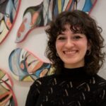 A person with brown curly shoulder length hair wearing a black sweater standing in front of a painting.