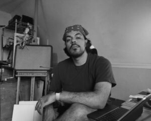 Black and white image of Orlando Caraballo Ortiz wearing a patterned bandana and gray t-shirt sitting in an arts studio.