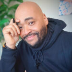 Bald man with a beard smiles warmly, resting his hand on his forehead. He's wearing a dark hoodie, with a blurred green background.