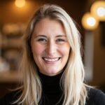 Smiling woman with long blonde hair in a black turtleneck inside a cozy cafe with warm lighting. The ambiance is inviting and cheerful.