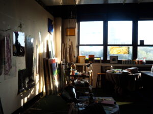 Sunlit EAR artist’s studio with paintings, canvases, and art supplies scattered throughout; large windows let in natural light.