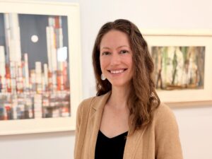 A woman smile at the camera with brown curly hair. Stands infront of art work wearing a black shirt and tan sweater.