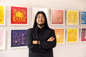 A Chinese man will long black hair, smiling with his arms cross his chest. Colorful frames art illustration lining the back wall